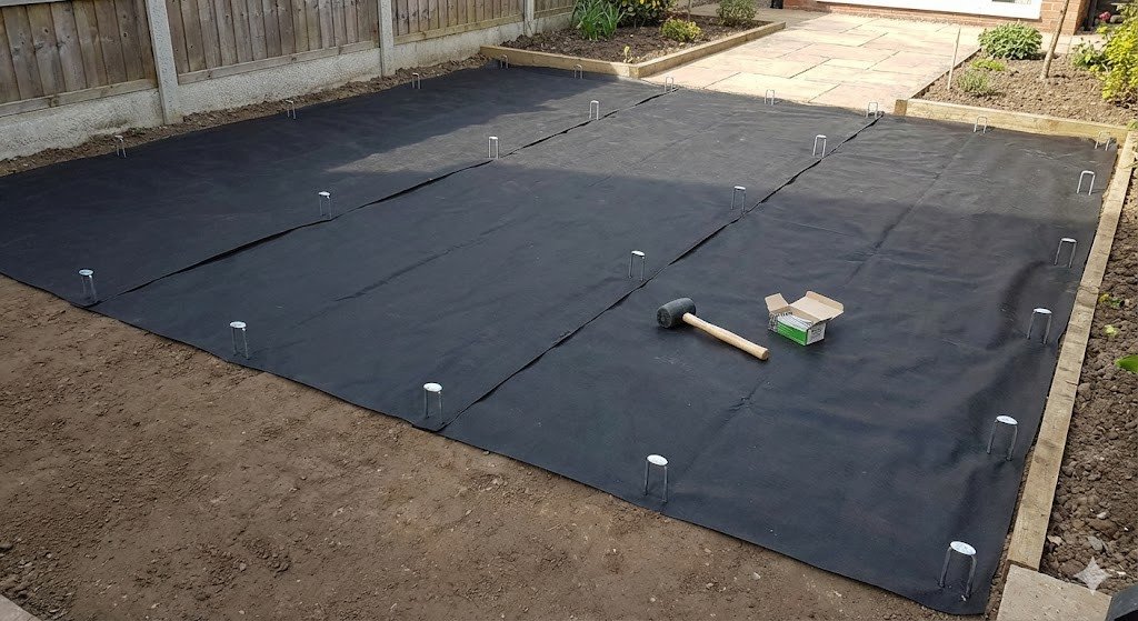 black weed membrane laid flat over a prepared ground base. The membrane is neatly spread with edges overlapping, and ground pins are visible securing it in place. 